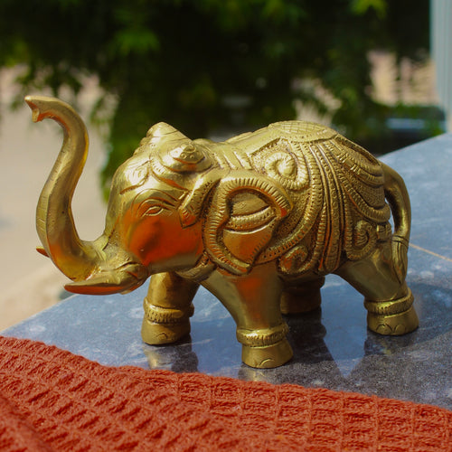 A decorative brass elephant figurine with intricate engravings, standing on a dark stone surface with a blurred outdoor background and a red textured fabric in the foreground.