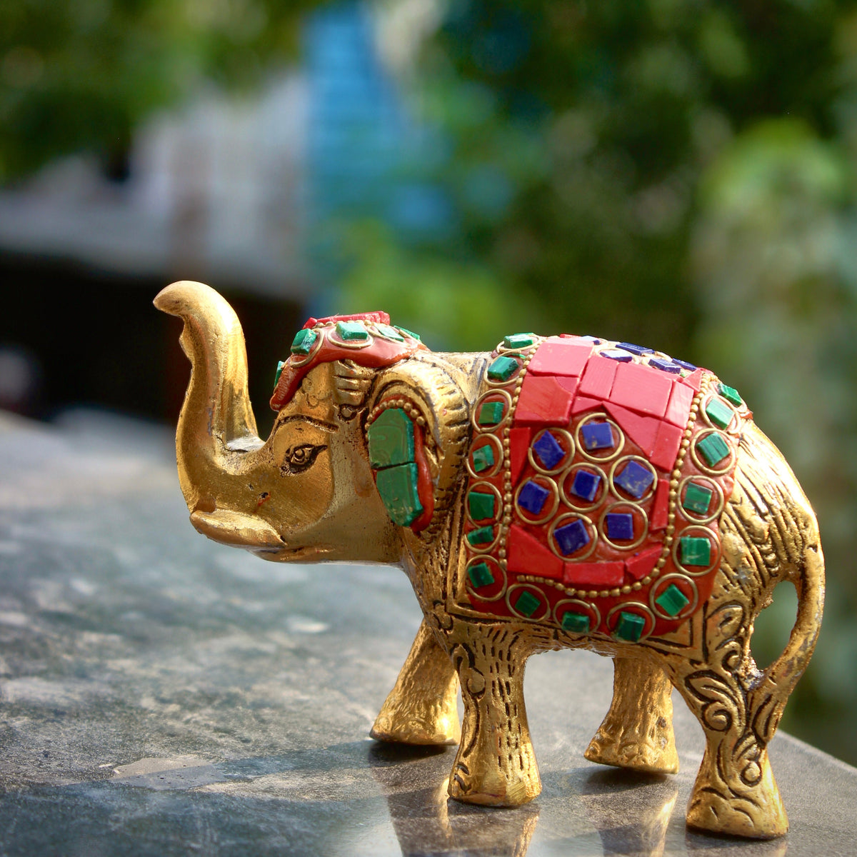 A decorative gold-toned elephant figurine with a red saddle and green and blue gem-like accents, standing on a dark reflective surface with a blurred green outdoor background.