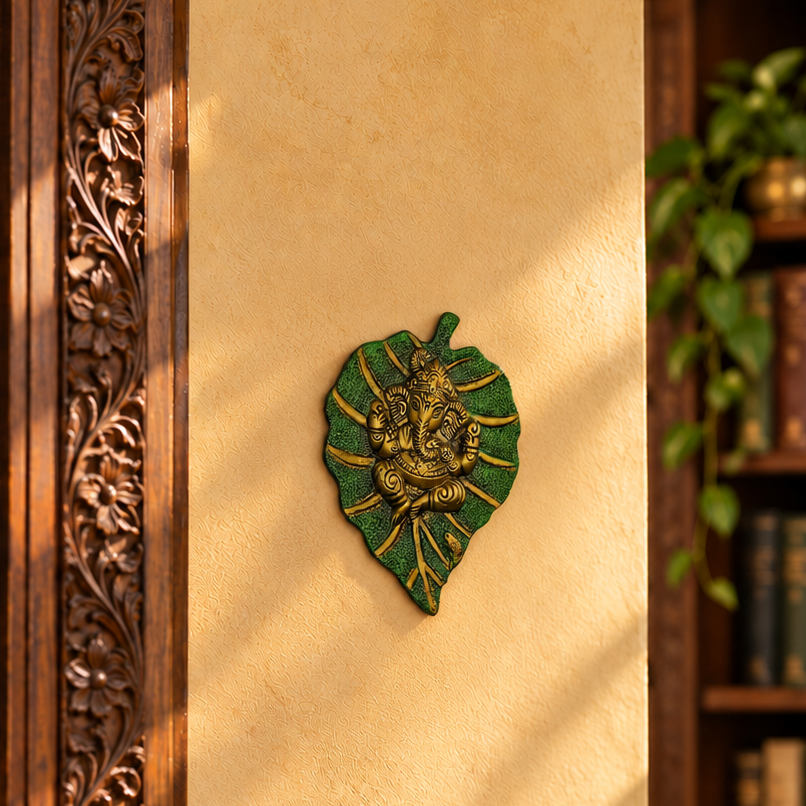 A decorative wall plaque shaped like a green leaf with a gold-toned seated deity figure in the center, mounted on a warm beige wall. The scene includes a carved wooden panel on the left and a blurred bookshelf with hanging greenery on the right.