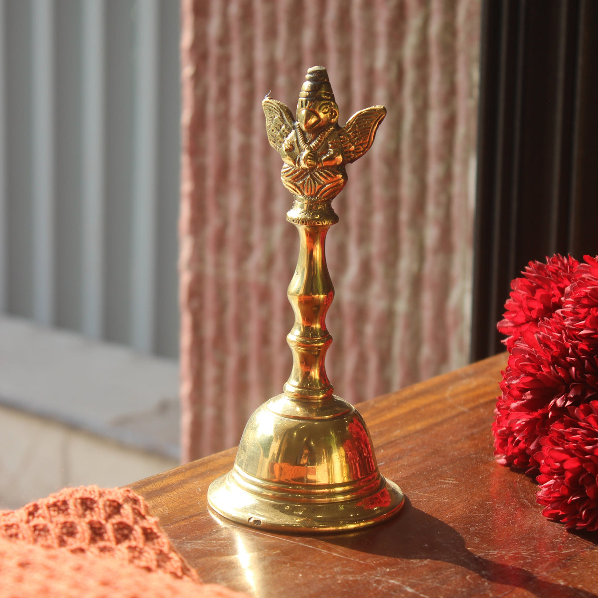 A polished brass bell with a decorative deity figurine on top, resting on a wooden table next to red flowers and a textured orange knit fabric.